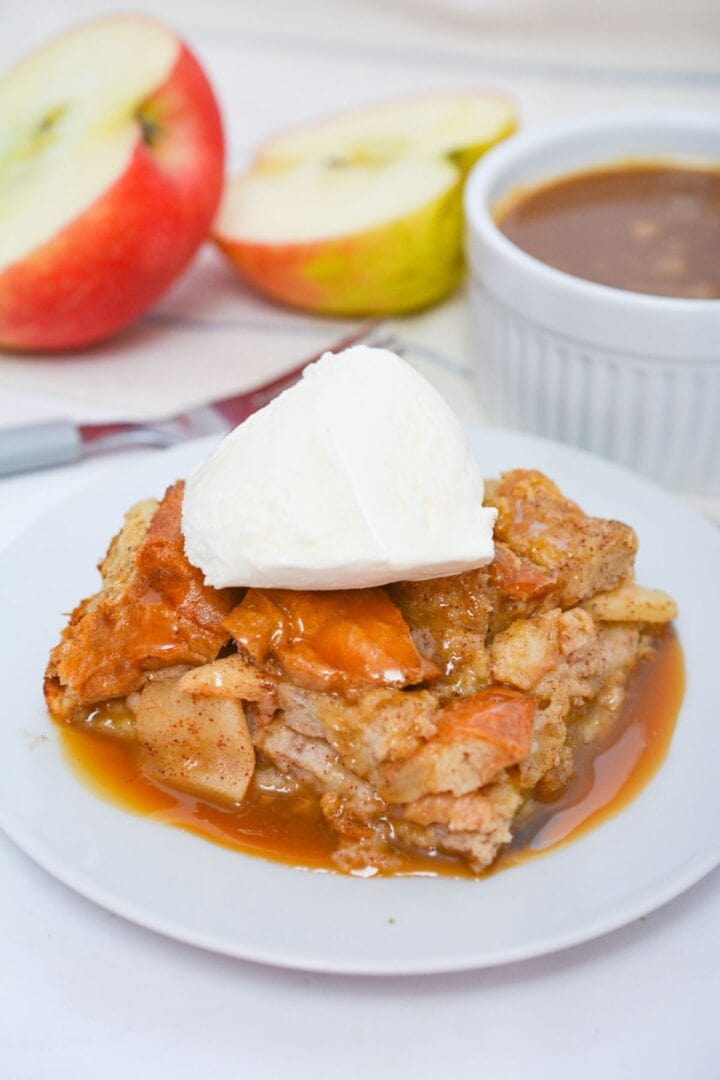 A slice of apple bread pudding with bourbon sauce is topped with vanilla ice cream on a white plate, accompanied by caramel sauce, fresh apple slices, and a knife in the background.
