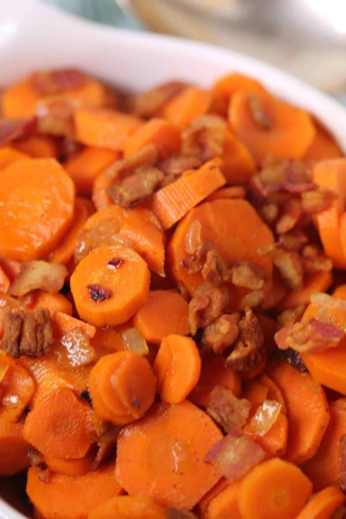 A dish of sliced cooked carrots glazed with brown sugar, mixed with small pieces of bacon, served in a white bowl.