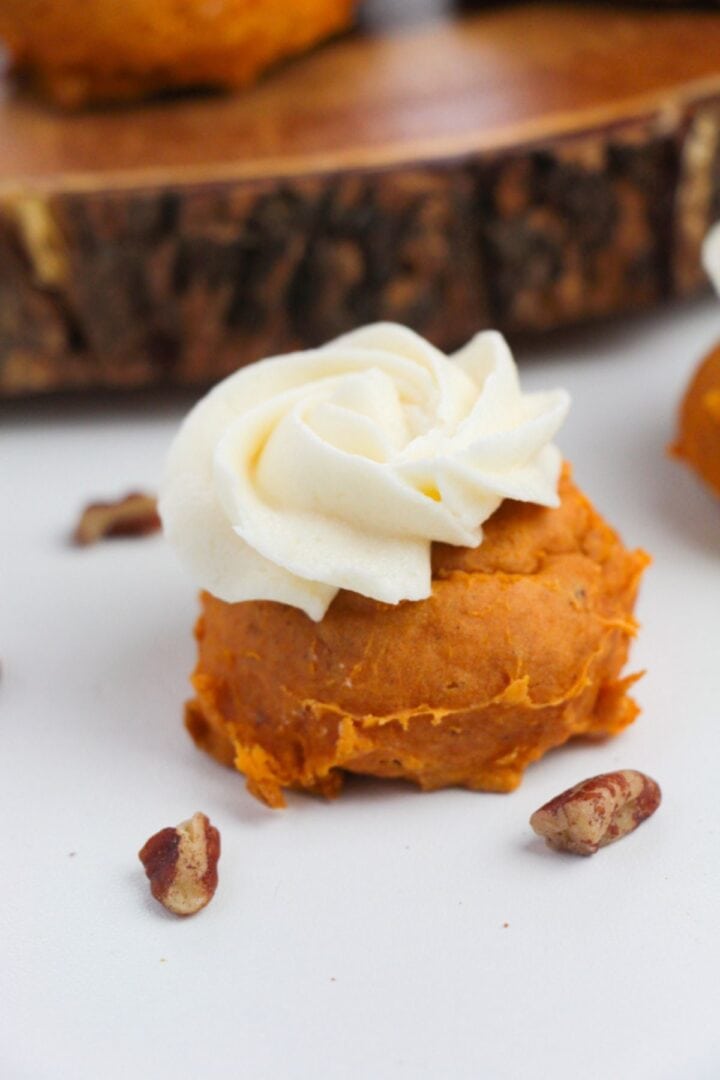 A pumpkin-flavored dessert made with Cake Mix Pumpkin Spice Cookies, topped with a swirl of white frosting and placed on a white surface with pecan pieces nearby.