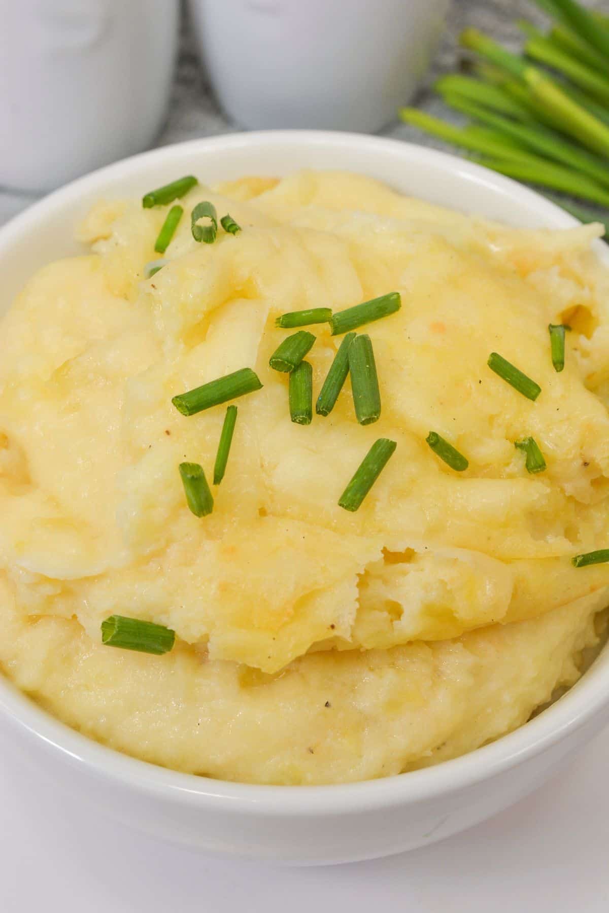 A white bowl filled with cheesy mashed potatoes, garnished with chopped chives, placed on a white surface.