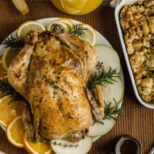 Roast chicken garnished with orange, apple, and rosemary on a plate, next to a baking dish of stuffing. A pitcher of orange juice and wooden utensils rest on a brown placemat—perfect with Citrus Turkey Brine flavors.