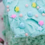 A slice of Cotton Candy Cake with blue frosting, adorned with pastel pink and blue round sprinkles and yellow and pink star sprinkles, rests elegantly on a floral plate.