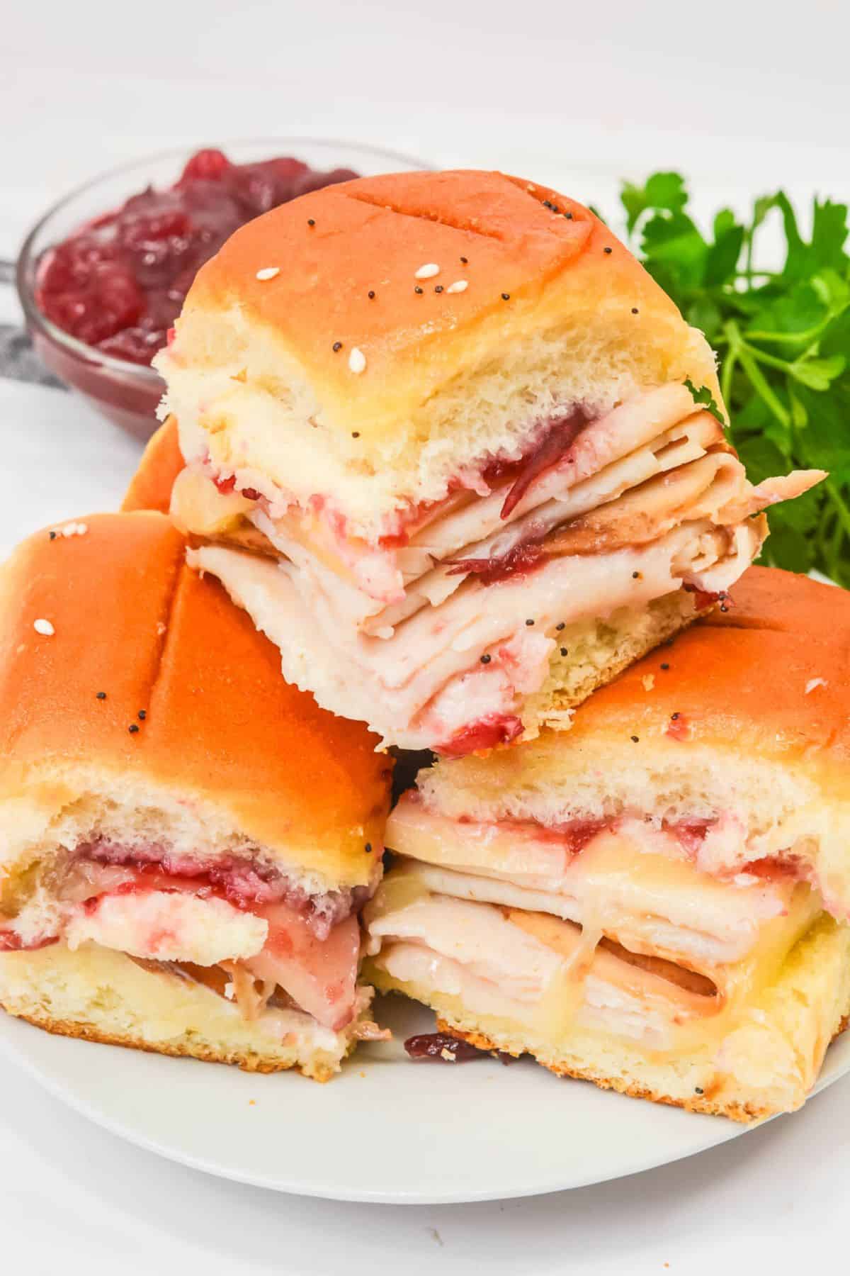 Three Cranberry Turkey Sliders with cheese and cranberry sauce are stacked on a white plate, garnished with parsley, with a bowl of cranberry sauce in the background.
