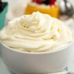 A white bowl filled with swirled cream cheese whipped cream sits on a light surface, with a blurred dessert and spoon in the background.