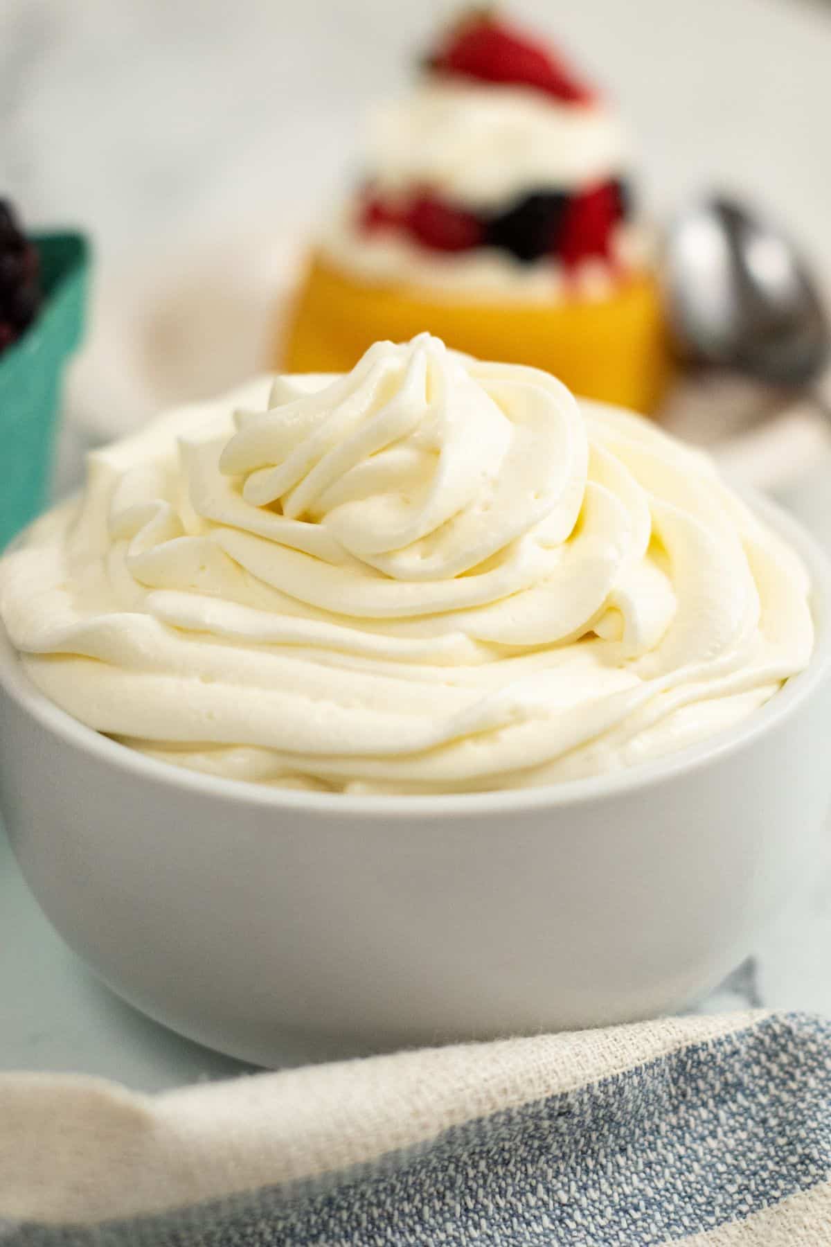 A white bowl filled with swirled cream cheese whipped cream sits on a light surface, with a blurred dessert and spoon in the background.