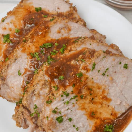 Slices of roasted crock pot pork tenderloin topped with sauce and garnished with herbs on a white plate. Nearby is a small bowl containing additional sauce.