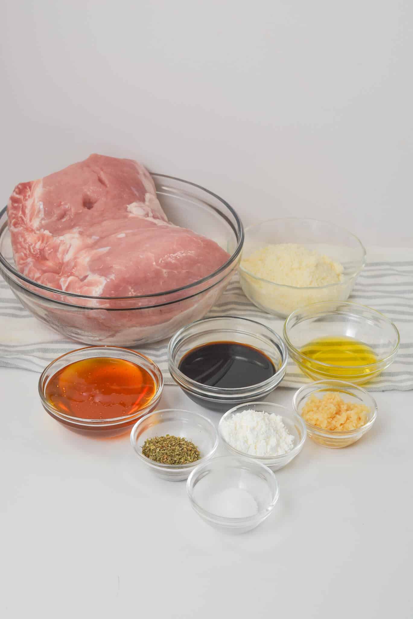 Raw pork tenderloin in a glass bowl is surrounded by small bowls of spices, grated cheese, oil, and soy sauce on a striped cloth, all ready to be transformed into a succulent crock pot masterpiece.