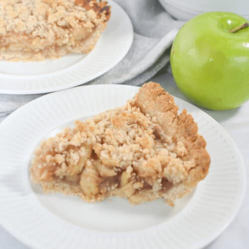 A slice of easy apple crumble pie on a white plate. Another slice of pie and a bowl of green apples are in the background.