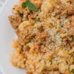 Close-up of a plate of creamy sausage risotto with chunks of meat, topped with grated cheese and garnished with fresh herbs.