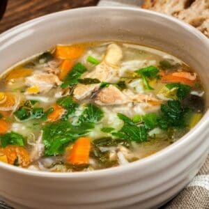 A bowl of Turkey Rice Soup with pieces of turkey, carrots, celery, rice, and fresh herbs, served with a slice of bread in the background.