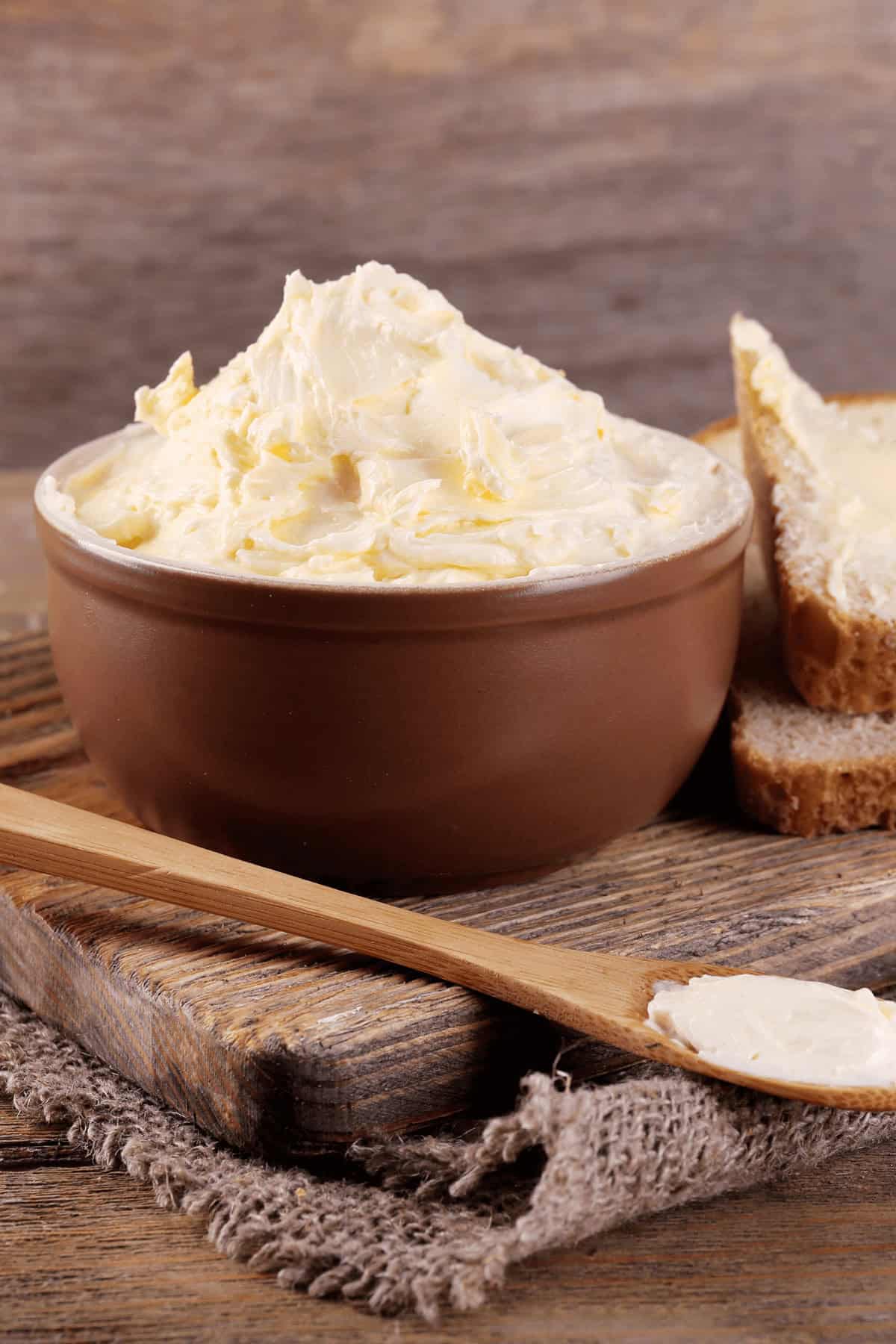 A bowl of homemade butter sits on a wooden board next to a buttered slice of bread and a wooden knife.