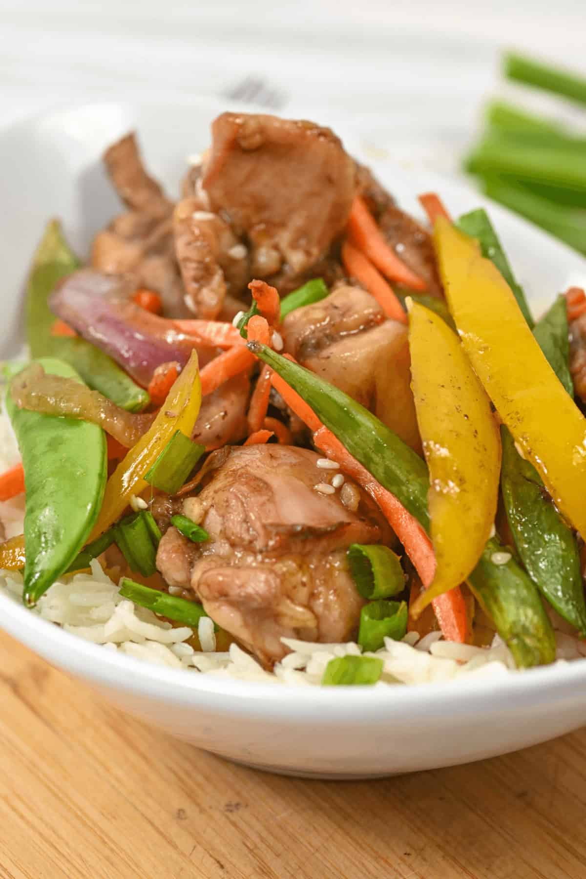 A bowl of rice topped with honey sesame chicken pieces, snow peas, carrots, yellow bell peppers, and red onions.