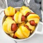 A white dish filled with Mini Pigs in a Blanket—savory mini sausages wrapped in golden, baked crescent roll dough—rests on a marble surface beside a gray and white striped cloth.