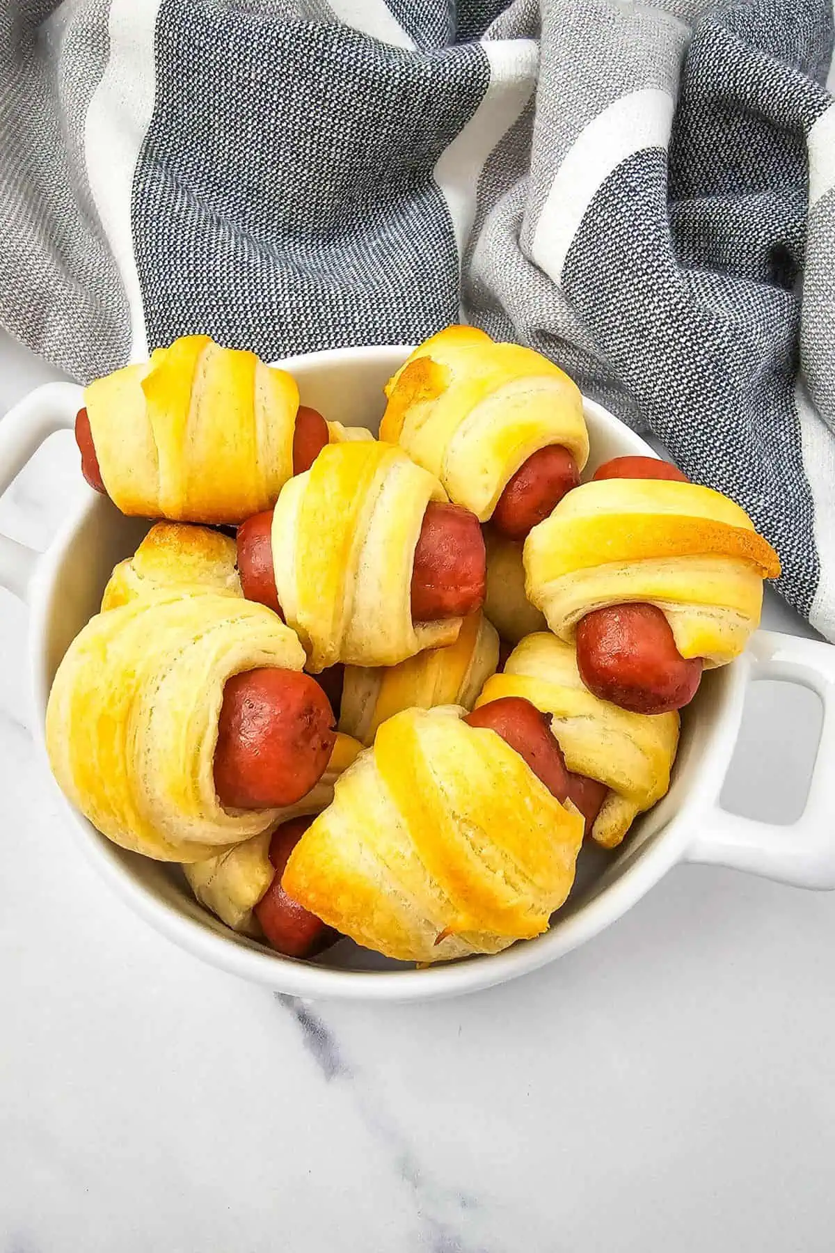 A white dish filled with Mini Pigs in a Blanket—savory mini sausages wrapped in golden, baked crescent roll dough—rests on a marble surface beside a gray and white striped cloth.
