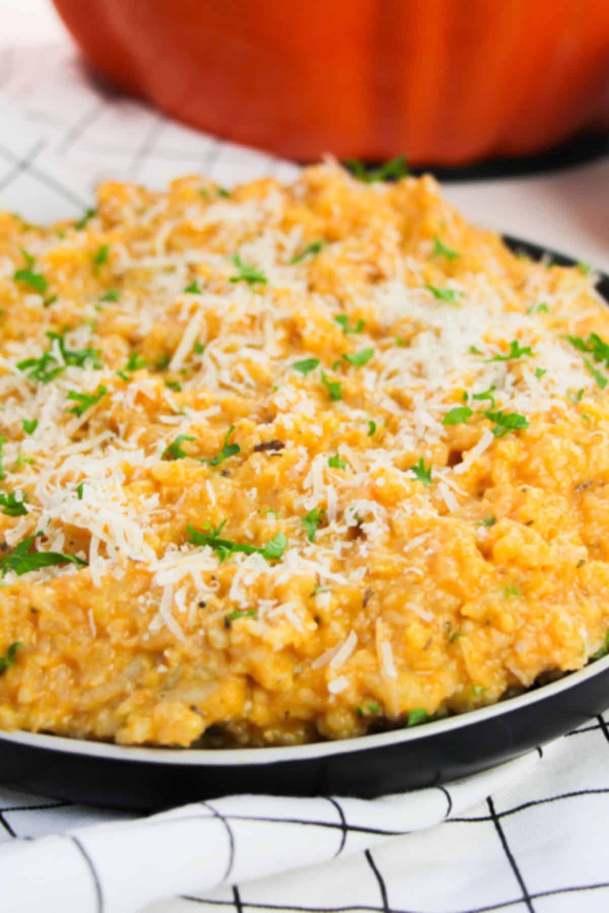 A plate of creamy pumpkin risotto topped with grated cheese and chopped herbs, placed on a white checkered cloth.
