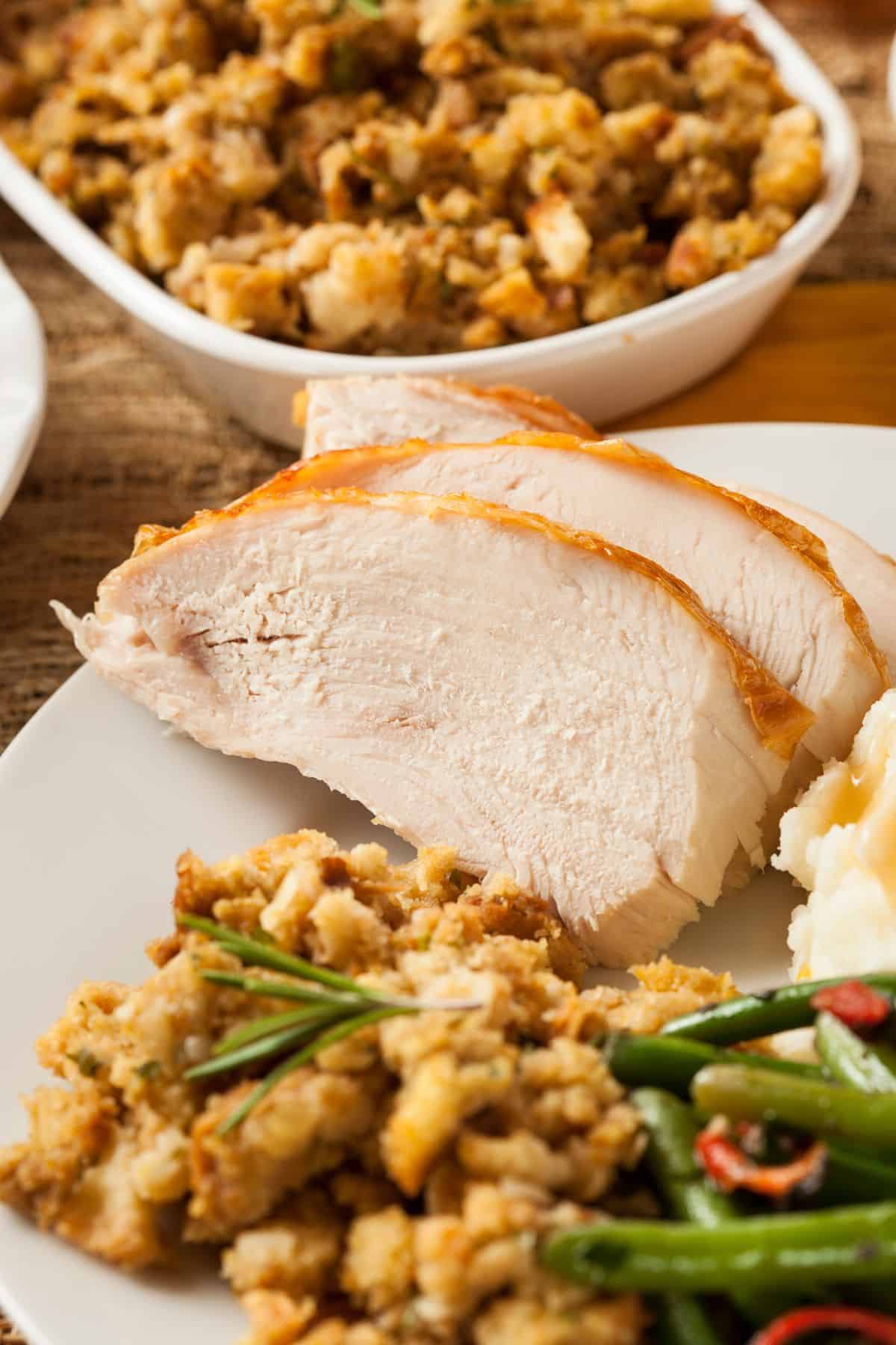 Slow Roasted Turkey Breast served with stuffing, green beans, and mashed potatoes on a white plate. A dish of extra stuffing is in the background.