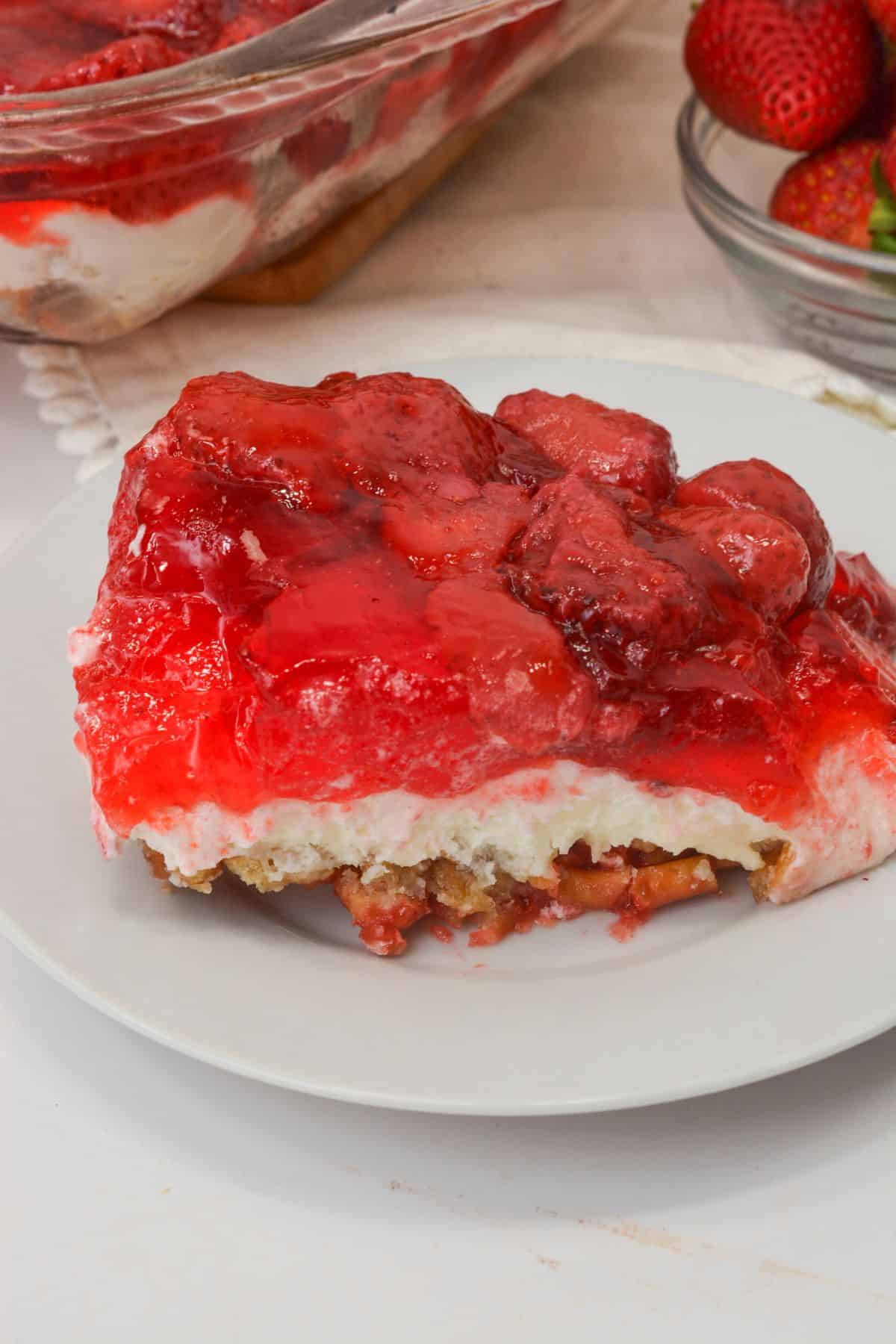 A slice of Pretzel Jello Salad dessert with a crumbly base, creamy middle layer, and a topping of glazed strawberries sits elegantly on a white plate.