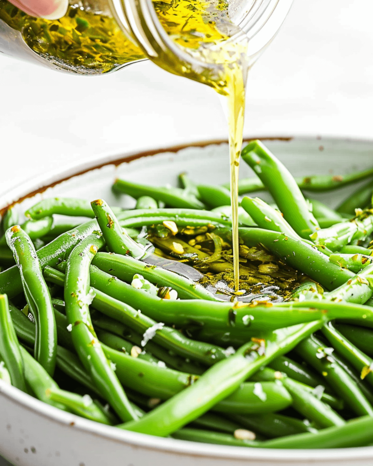 A person pouring Italian olive oil over green beans in a bowl.